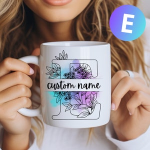May include: White ceramic mug with a black outline of the letter "E" decorated with floral designs and watercolor accents. The text "custom name" is printed below the letter.