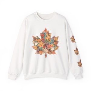 May include: A white crewneck sweatshirt featuring a large maple leaf graphic on the center chest composed of colorful puzzle pieces with autumn motifs including pumpkins, ghosts, and fall foliage. Three smaller matching leaf graphics appear on the sleeve.