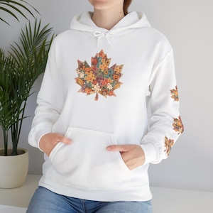 May include: A white hooded sweatshirt featuring a large maple leaf graphic on the chest and smaller matching leaf graphics on the sleeve. The leaf design is composed of a colorful puzzle piece pattern with autumn motifs including pumpkins and ghosts.