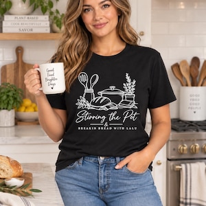 May include: A black short-sleeve t-shirt featuring a white graphic of kitchen utensils, a pot, bread, and a plant. The text reads Stirring the Pot & BREAKIN BREAD WITH LALU. A white mug with the text Good Food Brighter Days is held in one hand.