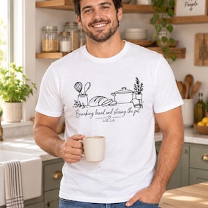 May include: A white short-sleeved t-shirt featuring a black line art graphic of kitchen utensils, a loaf of bread, and a pot. The text reads "Breaking bread and stirring the pot" with "With Lulu" underneath and a small heart symbol in between.