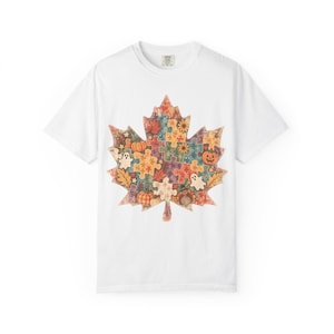 Puede incluir: Una camiseta blanca de manga corta con un gráfico grande de hoja de arce compuesto por piezas de rompecabezas coloridas. El diseño incluye motivos otoñales como calabazas, girasoles, fantasmas y un pavo integrados en el patrón.