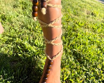 May include: Barefoot sandals made of natural rope, wrapped around the leg and foot. The sandals feature dark wooden beads as decorative accents. The foot is painted with pink nail polish, and the sandals are set against a backdrop of green grass.