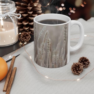 May include: White ceramic mug filled with a dark beverage. The mug features a wheat field design. The mug is on a light gray surface, with a lit candle in a glass jar, cinnamon sticks, and pine cones.