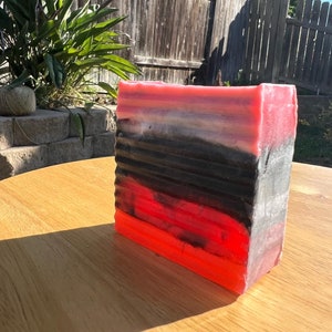 Pode incluir: Uma barra de sabão retangular com camadas alternadas de rosa, branco, preto e vermelho. O sabão está sobre uma mesa de madeira.