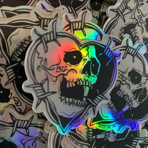 Könnte beinhalten: Ein Stapel irisierender holografischer Aufkleber mit einem schwarzen und weißen Totenkopfdesign mit Stacheldraht. Der Totenkopf hat die Form eines Herzens und hat ein einzelnes Auge.