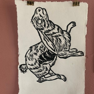 May include: Black and white linocut print of a rabbit with its head tilted back, looking up. The rabbit is depicted in a stylised, almost skeletal, manner.
