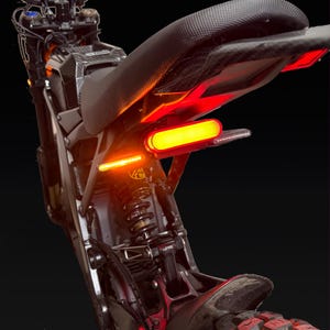 Peut inclure: Une section arrière de moto noire avec une selle texturée, un feu arrière horizontal rouge vif et des clignotants orange. Le système de suspension exposé est visible sous la selle, avec des accents style fibre de carbone sur le cadre.