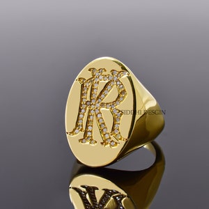 Puede incluir: Un anillo de sello de oro con una cara ovalada que presenta las letras "KR" engastadas con pequeños diamantes. El anillo está sobre una superficie negra.