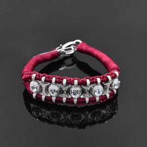May include: A maroon cord bracelet featuring a series of silver-toned metal links set with round, clear, faceted stones. The bracelet includes a silver-toned metal clasp and the text SIDDHI DESIGN is visible on the central metal link structure.