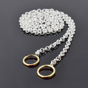 Puede incluir: Un collar de cadena de plata con dos anillos de oro en los extremos. La cadena está enrollada y los anillos cuelgan.