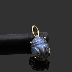 Puede incluir: Un colgante de escarabajo tallado, azul-grisáceo, con detalles dorados. El colgante tiene un lazo dorado para colgar y está sobre un fondo oscuro. El texto "SIDDHI DESIGN" es visible en el colgante.