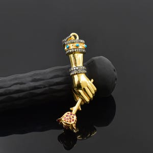 Op de afbeelding: Een goudkleurige hanger in de vorm van een hand die een roos vasthoudt. De hand is versierd met een diamantachtige band en turkooizen stenen. De roos heeft rode edelsteenaccenten. De hanger ligt op een zwarte ondergrond.
