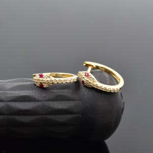 May include: A pair of gold-colored hoop earrings in a snake design. The earrings are encrusted with small, clear stones and feature red accents. The earrings are displayed on a black, textured surface.