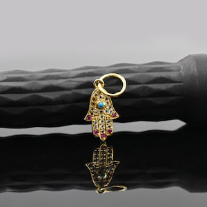 Op de afbeelding: Gouden Hamsa hand-hanger met een kleine turkooizen steen in het midden en rode en heldere stenen. De hanger wordt weerspiegeld op een glanzend zwart oppervlak.