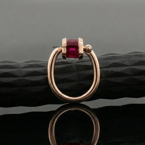Può includere: Un anello in oro rosa con una gemma rettangolare rosso intenso. L'anello ha un design unico con una fascia circolare e un elemento decorativo. L'anello è esposto su una superficie nera strutturata.