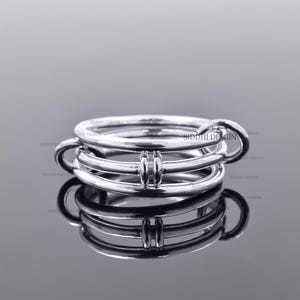 Puede incluir: Una pila de tres anillos plateados con un acabado pulido. Los anillos están entrelazados, con un elemento de diseño único en sus puntos de conexión. Los anillos se muestran sobre una superficie reflectante.