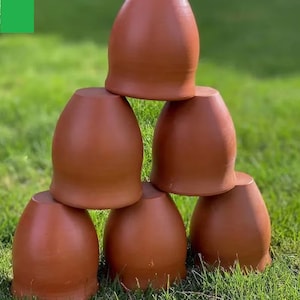 Może przedstawiać: Sześć glinianych kubków z terakoty ułożonych w kształt piramidy na zielonej trawie. Kubki są brązowo-czerwone i mają zaokrąglony kształt.