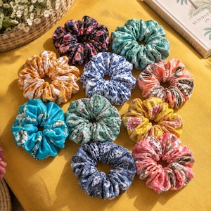 May include: A collection of ten fabric hair scrunchies in various colors and patterns, including floral prints in shades of blue, yellow, pink, green, and orange. Each scrunchie features a gathered, ruffled design with an elastic interior.