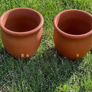 May include: Two terracotta flower pots, both round and unglazed, sitting on a bed of green grass.