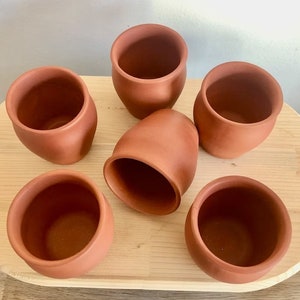 Puede incluir: Seis tazas de terracota en una bandeja de madera. Las tazas son todas del mismo tamaño y forma, y tienen un aspecto rústico y terroso.