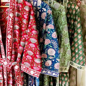 Op de afbeelding: Vier verschillende gepatroneerde gewaden hangen aan hangers. De gewaden zijn in rood, blauw, groen en bruin met bloemmotieven.