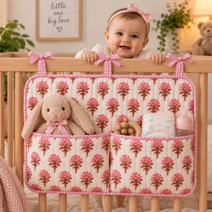 May include: A quilted fabric crib organizer featuring a repeating pink floral pattern on an off-white background with pink gingham trim and ties. The organizer has two large front pockets containing a plush bunny, a wooden rattle, diapers, and a bottle.