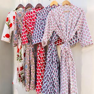Puede incluir: Cinco batas de algodón con estampado floral diferentes colgadas de perchas. Las batas son de varios colores, incluyendo blanco, rosa, rojo y azul. Las batas tienen un patrón floral sobre un fondo blanco. Las batas están todas atadas a la cintura.
