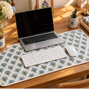 May include: A rectangular quilted desk mat featuring a repeating pattern of blue flowers with green stems on a white background. The mat has rounded corners and a white fabric border, providing a soft surface for a laptop, keyboard, and mouse.