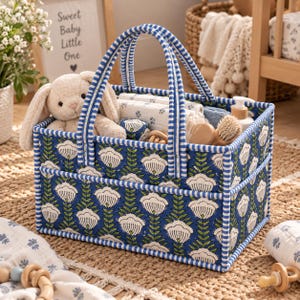 Puede incluir: Una cesta de almacenamiento rectangular de tela con un estampado floral azul y blanco, con asas y bordes a rayas azules y blancas. La cesta contiene un peluche de conejo beige, telas blancas estampadas plegadas y accesorios de madera.