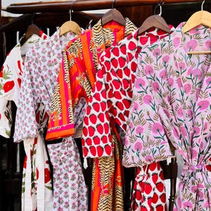 Op de afbeelding: Een verzameling kleurrijke badjassen die aan houten hangers hangen. De badjassen hebben verschillende patronen, waaronder grote rode klaprozen, roze bloemmotieven en heldere aardbeien. Eén badjas heeft een oranje en roze tijgerstreepmotief.