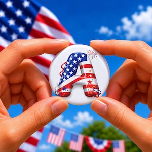 May include: A round white button featuring a large, three-dimensional letter A decorated with the American flag pattern of blue stars and red and white stripes. The letter is accented with small firework illustrations and red ribbons.
