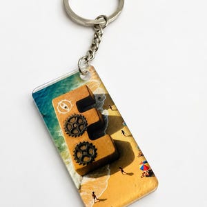 May include: A rectangular keyring featuring a beach scene with a wooden letter "E" decorated with gears. The keyring has a silver chain and ring. The bottom right corner of the keyring has the text "Ronnie Art".