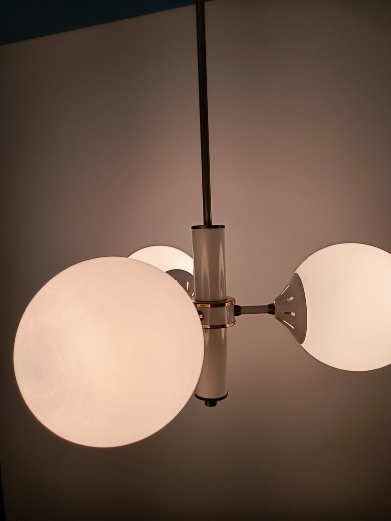 Peut inclure: Un lustre moderne avec trois abat-jours sph&eacute;riques en verre d&eacute;poli. Le luminaire a une finition blanche et dor&eacute;e avec une tige centrale et des bras s'&eacute;tendant pour tenir les globes. Le lustre est suspendu au plafond.
