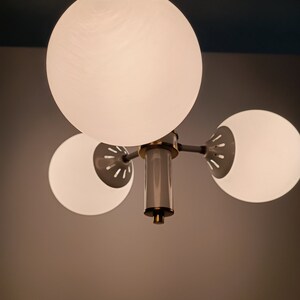 Peut inclure: Un lustre moderne avec trois abat-jours sph&eacute;riques en verre d&eacute;poli. Le luminaire a une finition en laiton et une colonne centrale avec un accent gris et dor&eacute;. Les abat-jours sont espac&eacute;s uniform&eacute;ment et &eacute;mettent une lumi&egrave;re douce et diffuse.