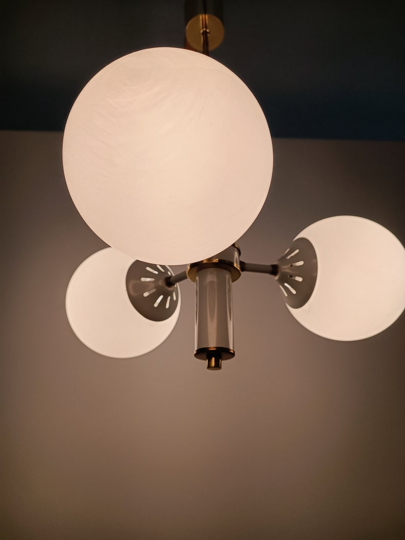 Peut inclure: Un lustre moderne avec trois abat-jours sph&eacute;riques en verre d&eacute;poli. Le luminaire a une finition en laiton et une colonne centrale avec un accent gris et dor&eacute;. Les abat-jours sont espac&eacute;s uniform&eacute;ment et &eacute;mettent une lumi&egrave;re douce et diffuse.