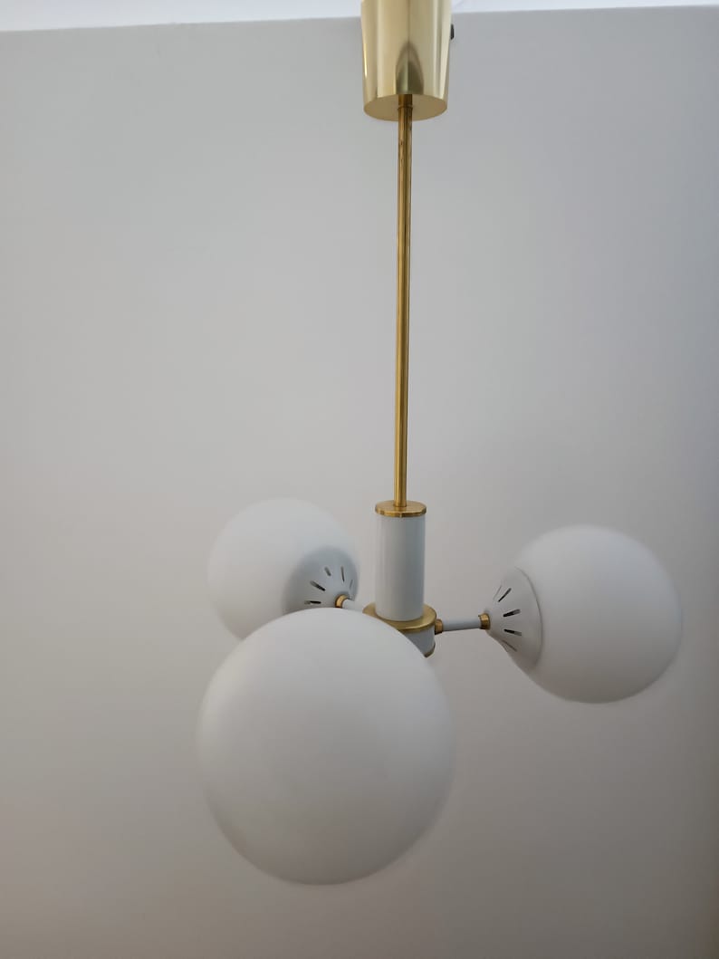 Peut inclure: Un plafonnier moderne avec trois abat-jours sph&eacute;riques en verre blanc. Le luminaire a une tige et des accents dor&eacute;s, avec un corps central blanc et dor&eacute;. La lumi&egrave;re est con&ccedil;ue pour &ecirc;tre mont&eacute;e au plafond.