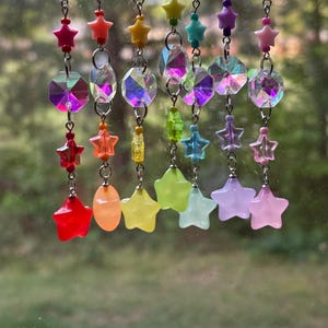 May include: A collection of rainbow-colored suncatchers hanging in front of a window. Each suncatcher features a chain with star-shaped and faceted crystal beads in various colors, including red, orange, yellow, green, blue, and purple.