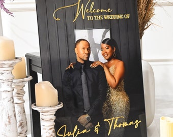 May include: Black welcome sign with gold lettering for a wedding. The sign says "Welcome to the wedding of Julia & Thomas March 21, 2024". A photo of the couple is in the center of the sign.