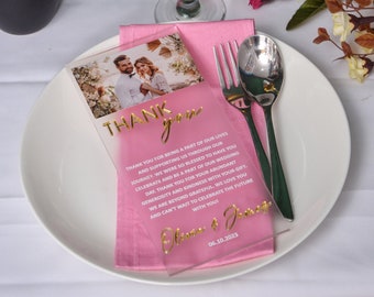 May include: Pink acrylic thank you card with gold lettering. The card features a photo of a couple and the text "THANK you" and "Thank you for being a part of our lives and supporting us through our journey. We were so blessed to have you celebrate and be a part of our wedding day. Thank you for your abundant generosity and kindness with your gift. We are beyond grateful. We love you and can't wait to celebrate the future with you!" and the date "06.10.2023".