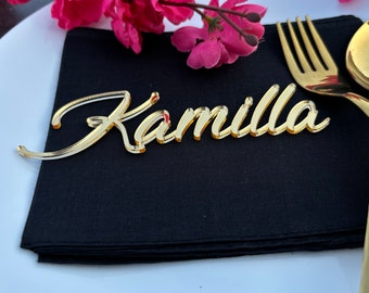 Puede incluir: Una etiqueta de nombre de acrílico dorado con el nombre Kamilla en letra cursiva. La etiqueta está colocada sobre una servilleta de tela negra junto a un tenedor de metal dorado y un plato blanco con flores rosas de fondo.