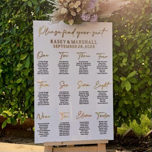 Peut inclure: Un plan de table de mariage blanc avec des lettres dorées. Le panneau indique "Veuillez trouver votre place" et répertorie les noms des invités sous les numéros un à douze. La date du mariage est le 28 septembre 2025.