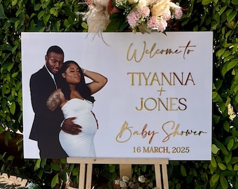May include: A white sign on an easel with the words "Welcome to Tiyanna + Jones Baby Shower 16 March, 2025" in gold lettering. A photo of a couple is on the left. The sign is decorated with flowers.