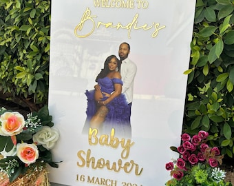 May include: White welcome sign with gold lettering for a baby shower. The sign says "Welcome to Brandie's Baby Shower 16 March 2024". A photo of a pregnant woman and a man is in the background.