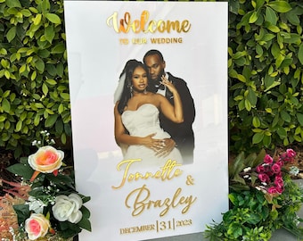 May include: White wedding sign with a photo of a couple in front of a pink and white background. The sign has gold lettering that says "Welcome to our wedding", "Tonnette & Brazley", and "December 31, 2023".