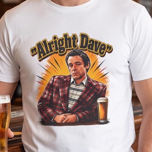 May include: A white t-shirt featuring a graphic illustration of a person in a red and black plaid blazer sitting at a table with a glass of beer. Above the figure, the text "Alright Dave" is printed in a bold, yellow, retro-style font.