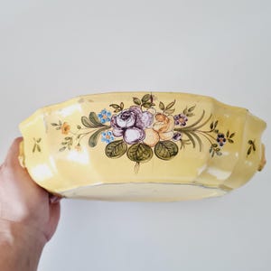 Puede incluir: Una fuente de cerámica amarilla con un diseño floral. Las flores pintadas a mano incluyen flores moradas, melocotón y azules con hojas verdes. La fuente tiene un borde festoneado y asas pequeñas a cada lado.