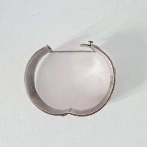 May include: A silver-toned metal hinged cuff bracelet with a smooth, polished interior surface. The bracelet features a secure clasp mechanism at the opening and includes a thin, delicate safety chain attached to both sides of the hinge for added security.