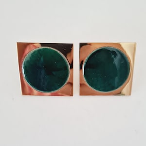 May include: A pair of square gold-toned cufflinks. Each cufflink features a circular, dark green enamel center. The cufflinks have a polished finish and a minimalist design, suitable for formal wear.