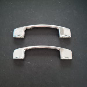 May include: Two shiny, silver cabinet pulls with a curved bar design. The pulls are rectangular at the ends and have a polished finish. They are set against a dark background.